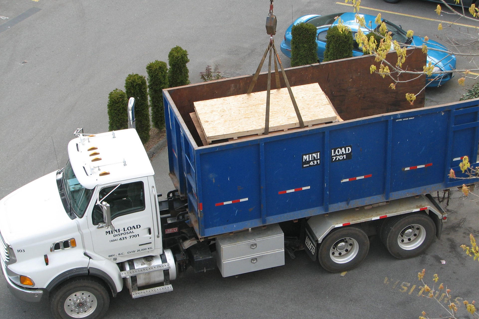 A large blue dump truck with a "Mini Load" logo is parked while a crane lowers a wooden crate into its container.