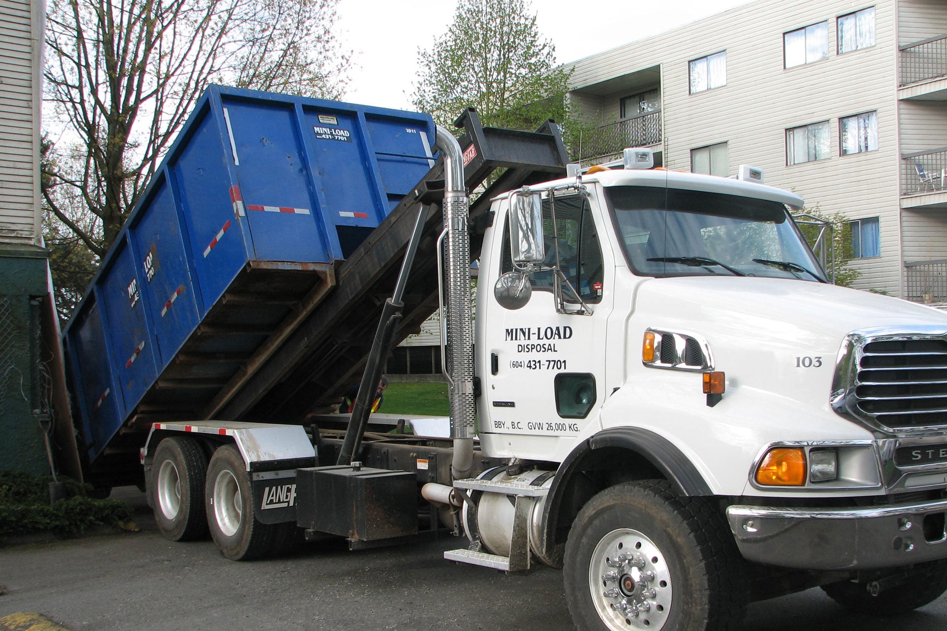 A white Mini-Load Disposal truck is unloading a large blue dumpster next to an apartment building.